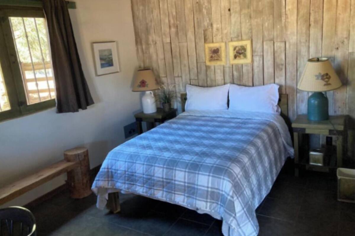 Quarto com cabeceira de madeira, cama com colcha xadrez, duas luminárias de cabeceira, uma mesinha com abajur e uma janela com cortinas.