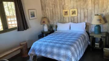 Un dormitorio con cabecera de madera, cama con colcha a cuadros, dos lámparas de noche, una mesita con lámpara y una ventana con cortinas.