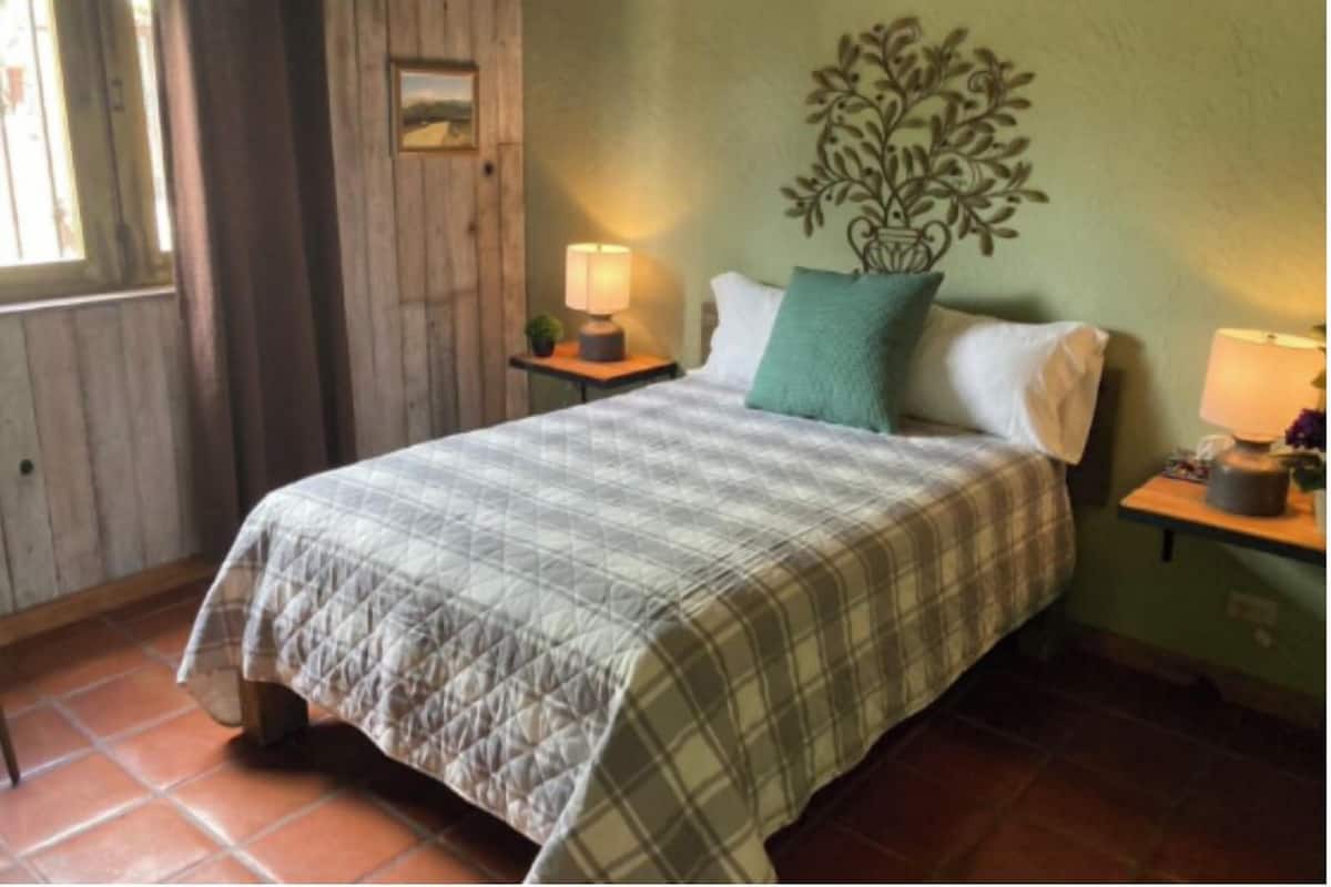 Quarto com cama, duas mesas de cabeceira, ar-condicionado fixado na parede e uma janela com cortinas.