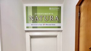 Un pasillo con una puerta que indica "APPARTAMENTO NATURE" y un felpudo con la palabra "Welcome".