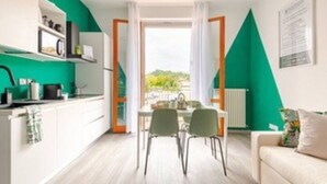 Una cocina moderna con armarios blancos y una pared de acento verde, un rincón de comedor con dos sillas y vistas al exterior con vegetación.