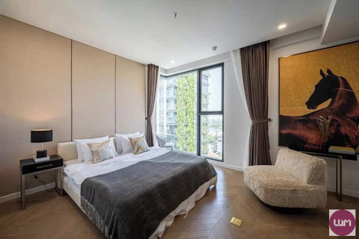 A modern bedroom with a large bed, a side table, a chair, and a large window with curtains.