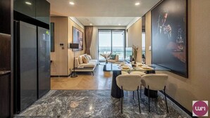 A modern hotel room with a dining area, a sofa, a TV, and a large window offering a city view.