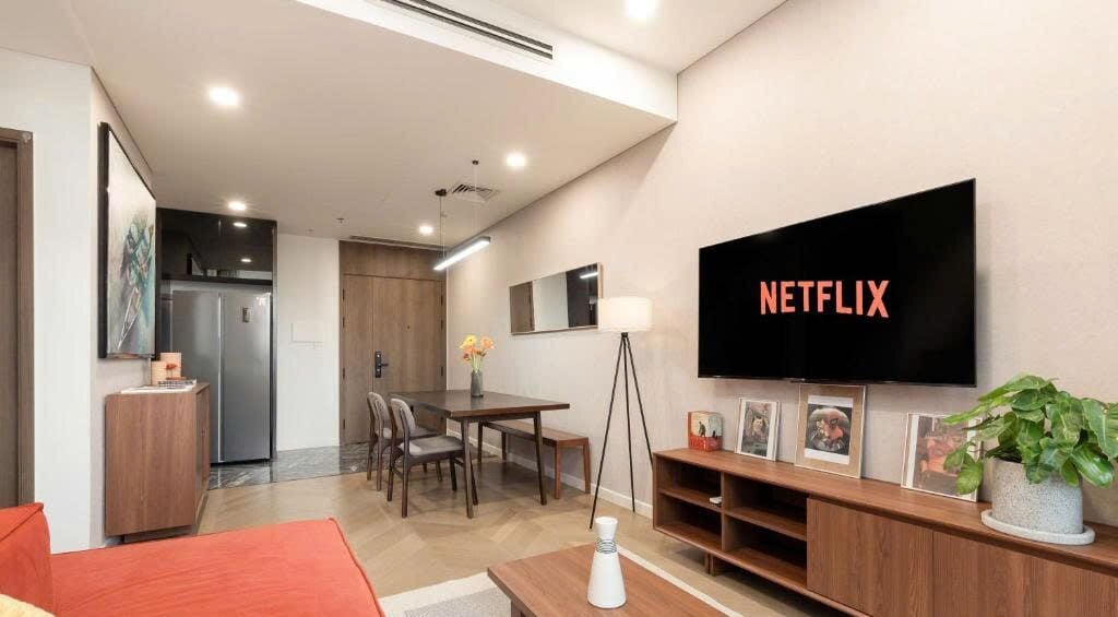 Een moderne woonkamer met een grote flatscreen televisie aan de muur, een eethoek met een houten tafel en stoelen, en een keuken met een koelkast en kastjes.