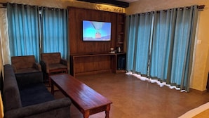 A living room with a sofa, two armchairs, a wooden coffee table, a television mounted on a wooden panel, and a small cabinet with a kettle.
