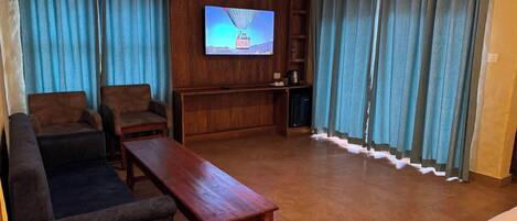 A living room with a sofa, two armchairs, a wooden coffee table, a television mounted on a wooden panel, and a small cabinet with a kettle.