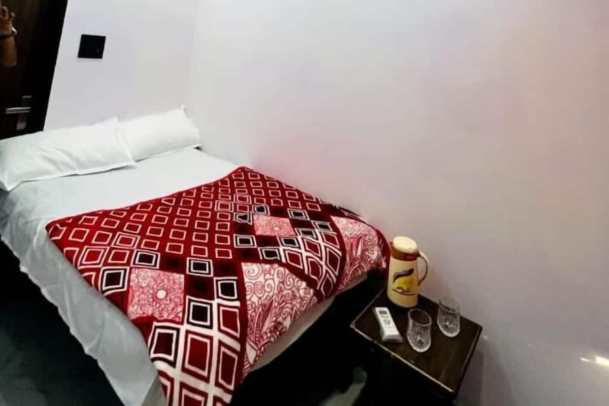 A hotel room with a bed, a ceiling fan, an air conditioning unit, a bedside table with a lamp, and a small table with a bottle and glasses.