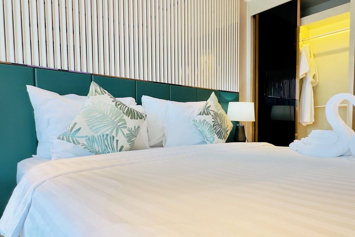 A neatly made bed with white linens and tropical leaf-patterned pillows, a bedside lamp, and a swan-shaped towel on the floor.