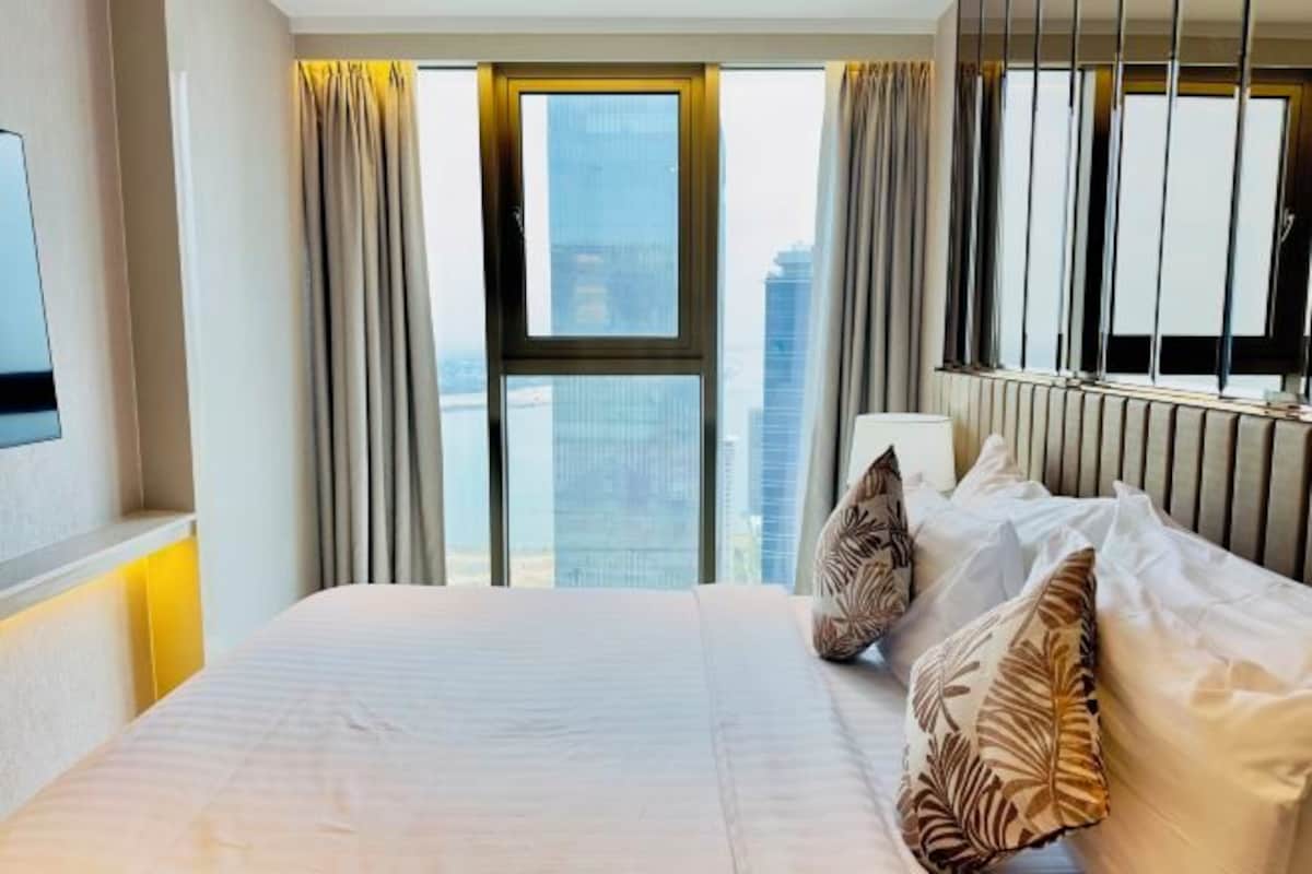 A hotel room with a large bed, a TV mounted on the wall, and a view of the cityscape through the window.