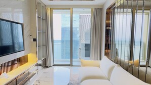 Una sala de estar moderna con una televisión de pantalla plana, una mesa de centro de mármol, un sofá y un amplio ventanal corredero que da a un balcón con vistas a la ciudad.