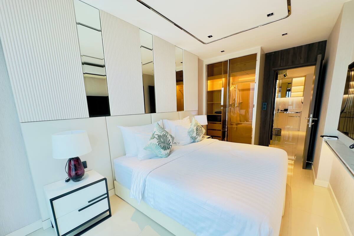 A modern hotel room with a large bed, a bedside table with a lamp, a wardrobe, and a mirror.
