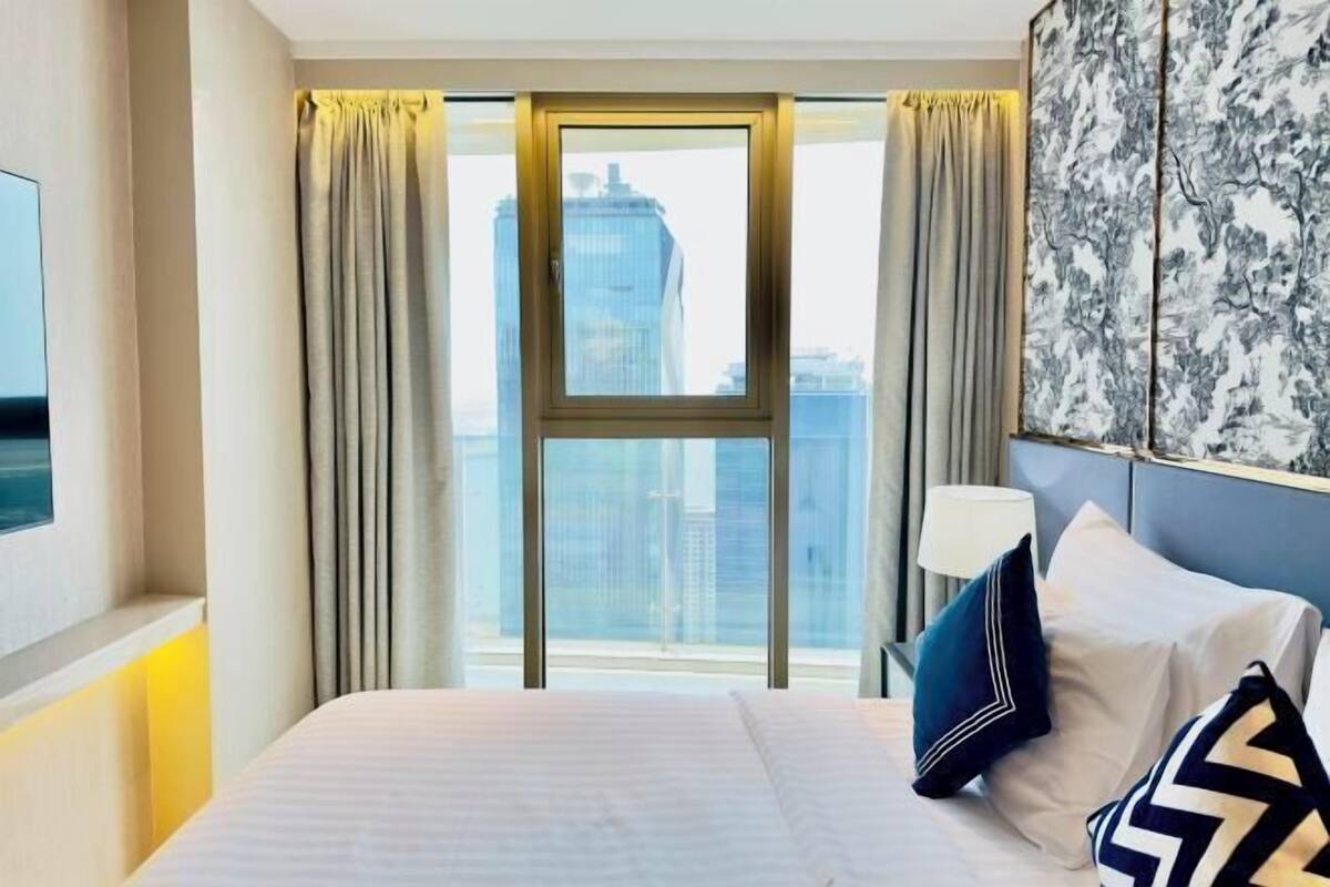 A hotel room with a large bed, a view of a cityscape, a window with curtains, and a wall with a patterned artwork.