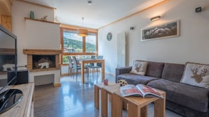 Uma sala de estar com lareira, um sofá com almofadas em formato de cabeça de veado, uma área de jantar com mesa e cadeiras e uma televisão sobre um suporte de madeira.