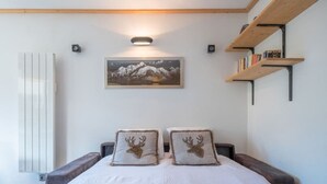 Quarto com cama, duas almofadas com estampas de veados, prateleira na parede com livros, quadro com paisagem de montanhas e radiador.