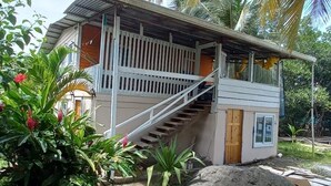 A single-story building with a wooden staircase leading to a porch, surrounded by lush greenery and palm trees.