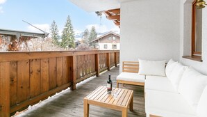 A wooden deck with a wooden table and white sofa, a bottle of wine, and a view of houses and trees. - Spacious apartment in the center, garden & sauna (Kirchberg in Tirol)