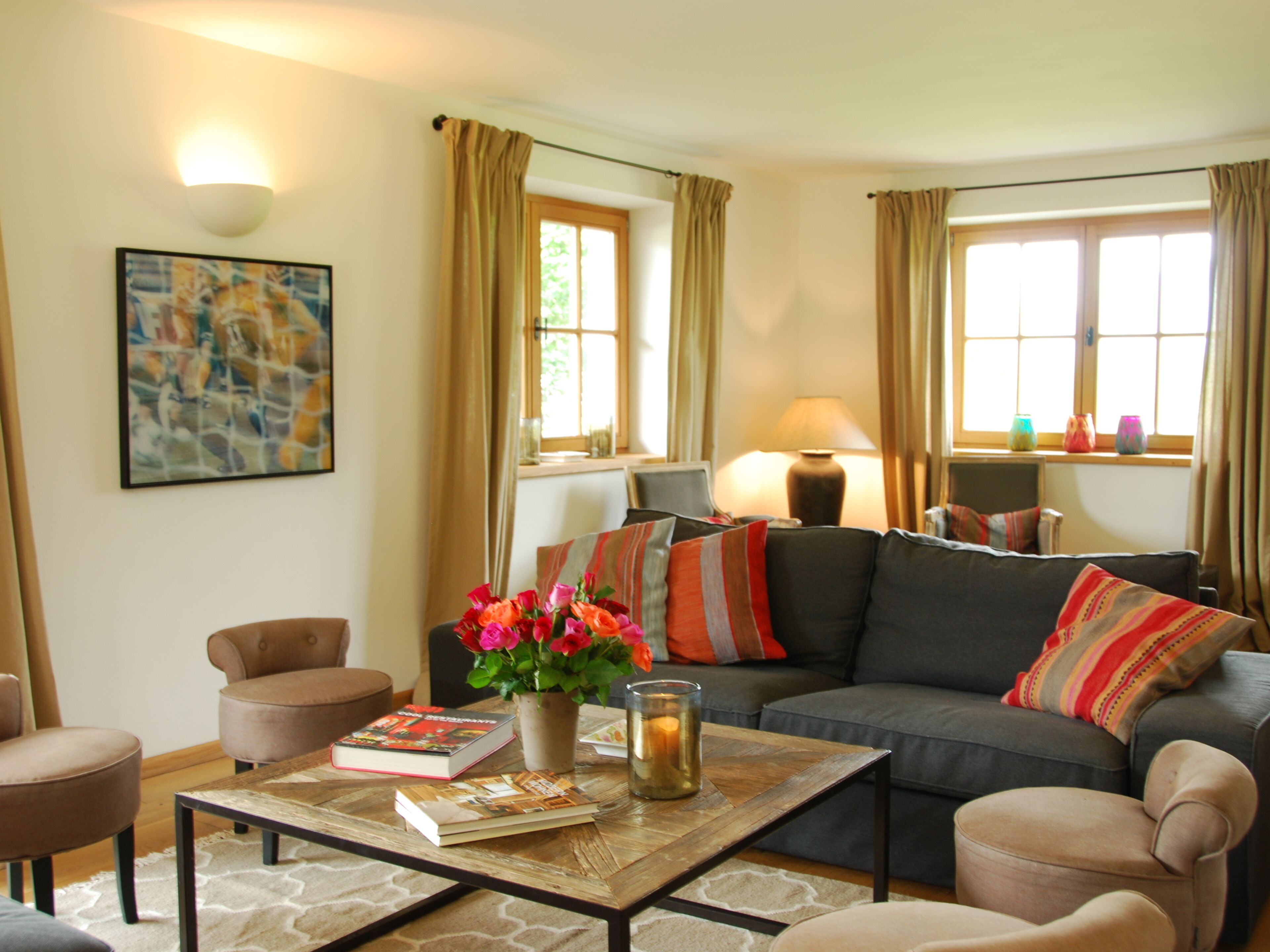 A living room with a sofa, armchairs, a coffee table with books and a vase of flowers, a painting on the wall, and a lamp.