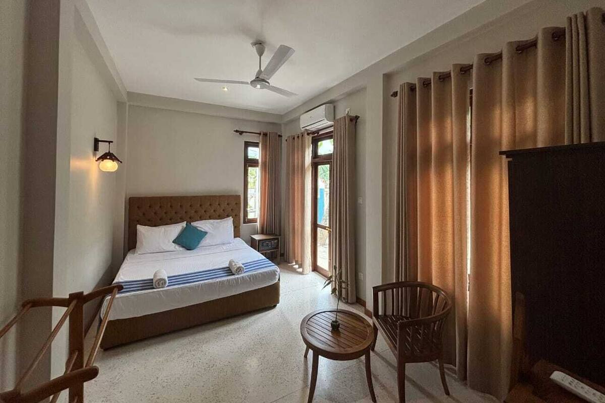 Una camera d'albergo con un letto, una sedia, un tavolo, una ventola a soffitto e tende.