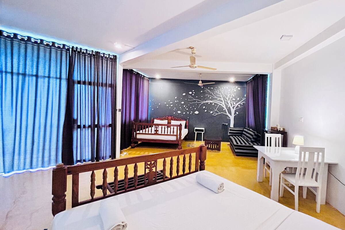 Una camera d'albergo con un letto, una scrivania con sedie, un divano, una ventola a soffitto e un murale raffigurante un albero.