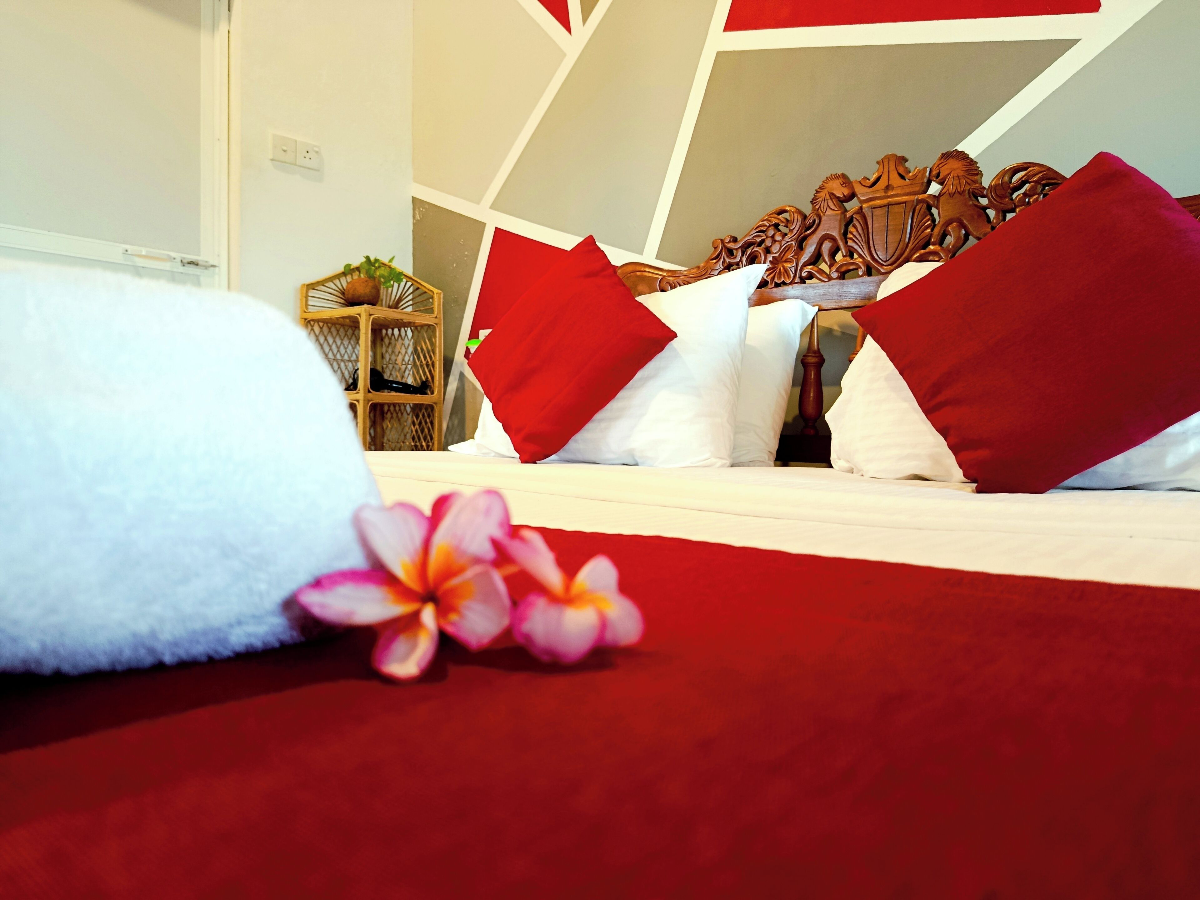 A neatly made bed with white linens and red pillows, a wooden headboard with a carved design, and a small plant on a shelf in the background.