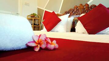 A neatly made bed with white linens and red pillows, a wooden headboard with a carved design, and a small plant on a shelf in the background.