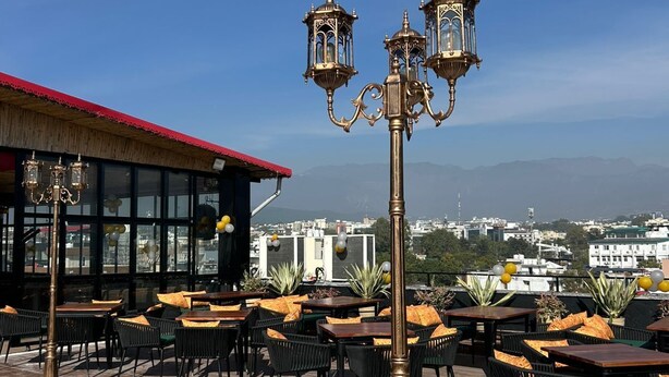 An outdoor seating area with tables and chairs, a decorative lamp post, and a view of the cityscape in the background. - HSR Hotels & Resorts (Dehradun)