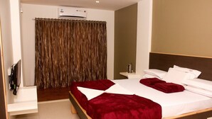 A hotel room with a large bed, a bedside table, a TV, and an air conditioning unit. - Hotel Anugraha Deluxe (Kolhapur)