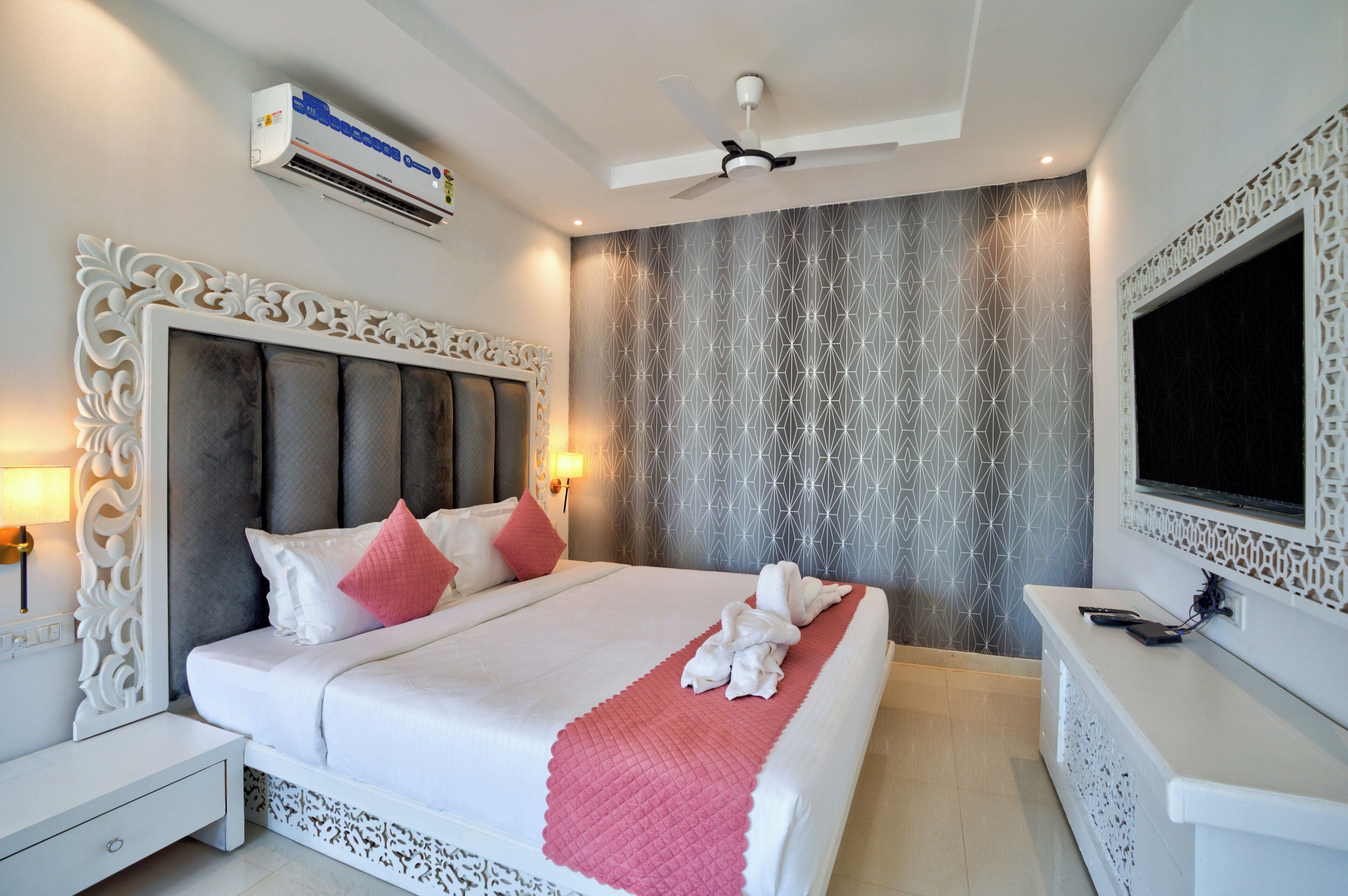 A neatly made bed with a white and pink bedspread, a decorative headboard, and a wall-mounted TV.