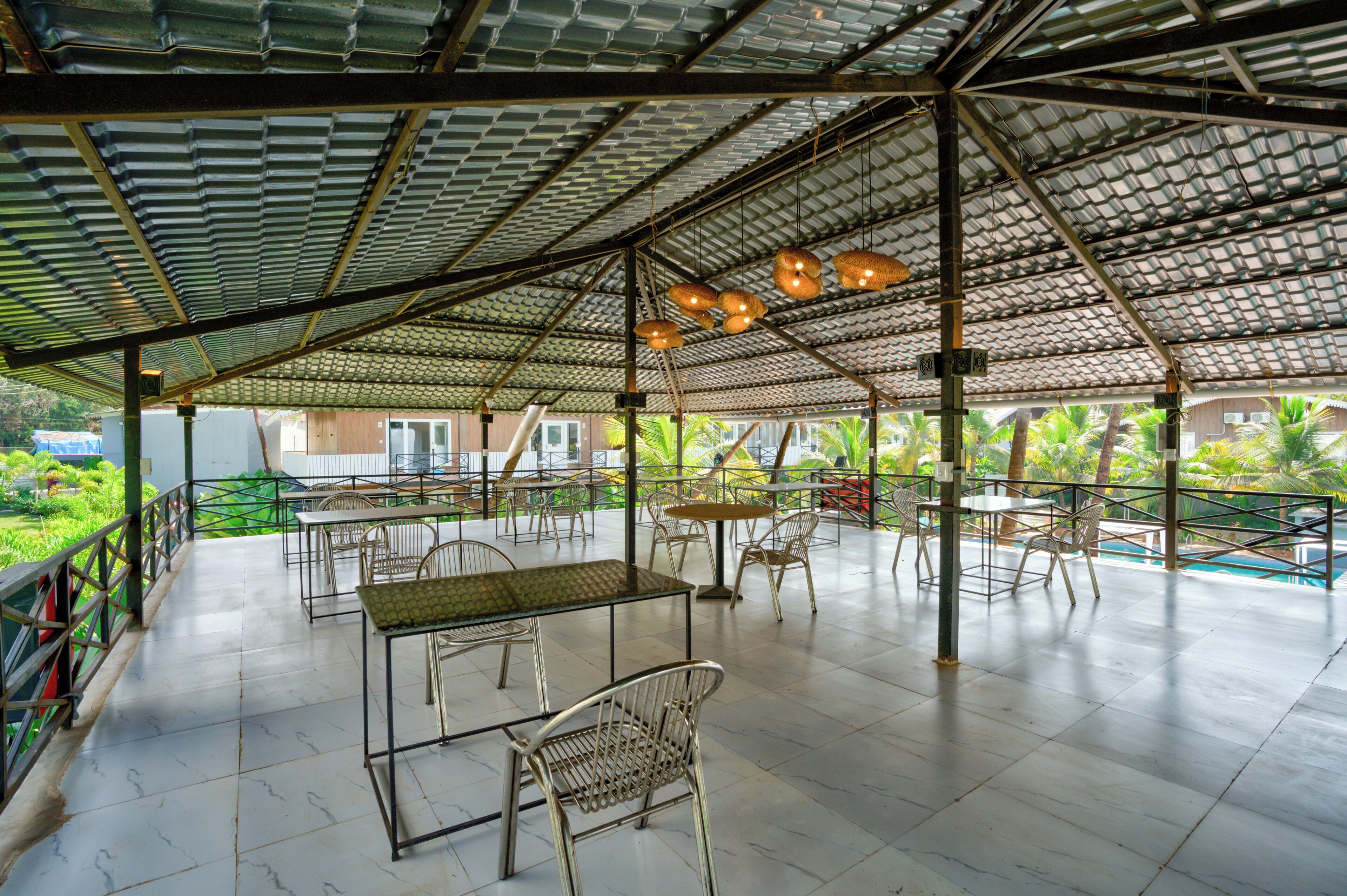 An outdoor patio area with metal chairs and tables, a corrugated metal roof, and a view of a pool and greenery.