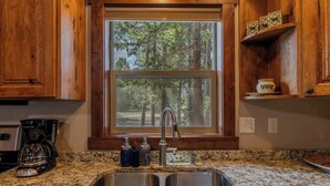 A kitchen with granite countertops, stainless steel sink, wooden cabinets, and a window with a forest view. - Burnt Hole (West Yellowstone)