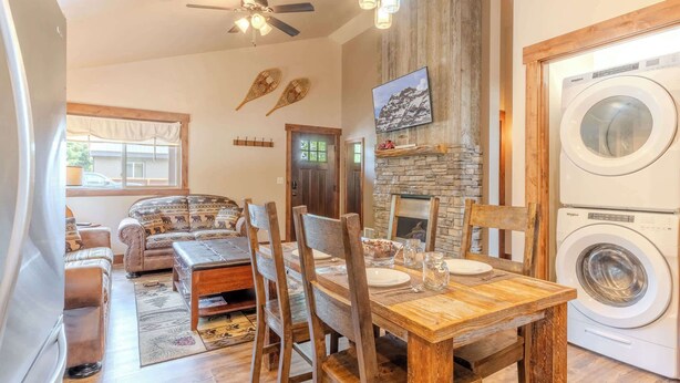 A cozy living room with a fireplace, a dining area with wooden furniture, and a laundry room with a front-loading washer and dryer. - Burnt Hole (West Yellowstone)