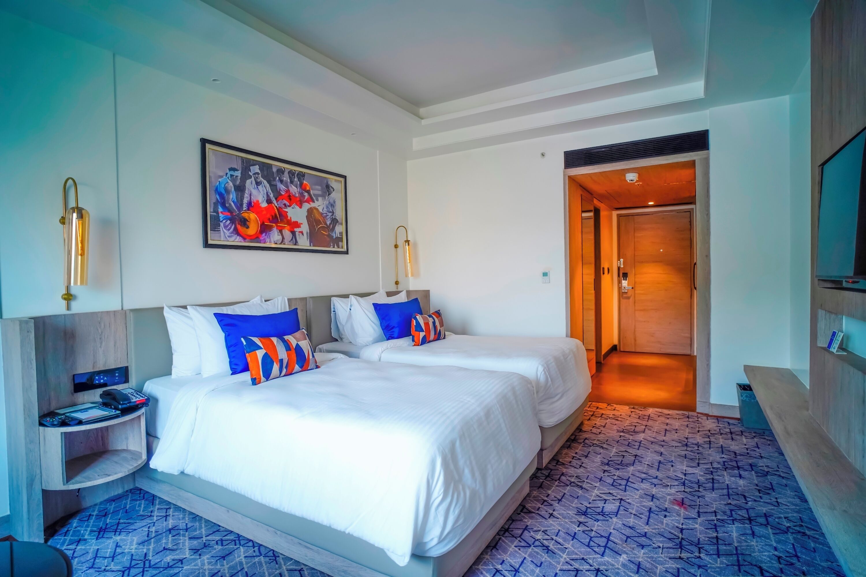 Habitación de hotel con dos camas, un cuadro en la pared, un escritorio con teléfono y cargador, y una alfombra estampada.