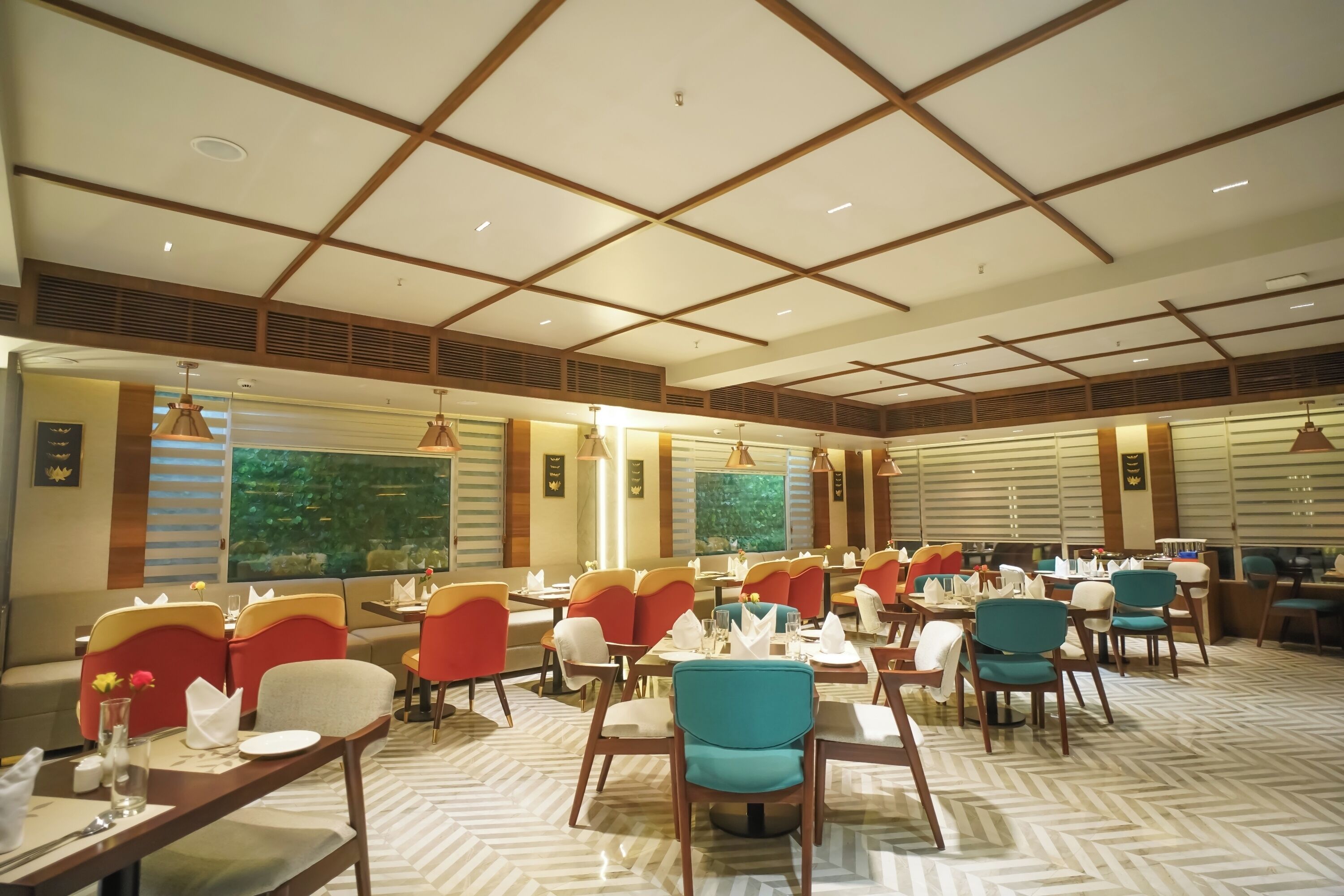 A spacious dining area with multiple tables and chairs, featuring a modern ceiling design and large windows with a view of greenery.
