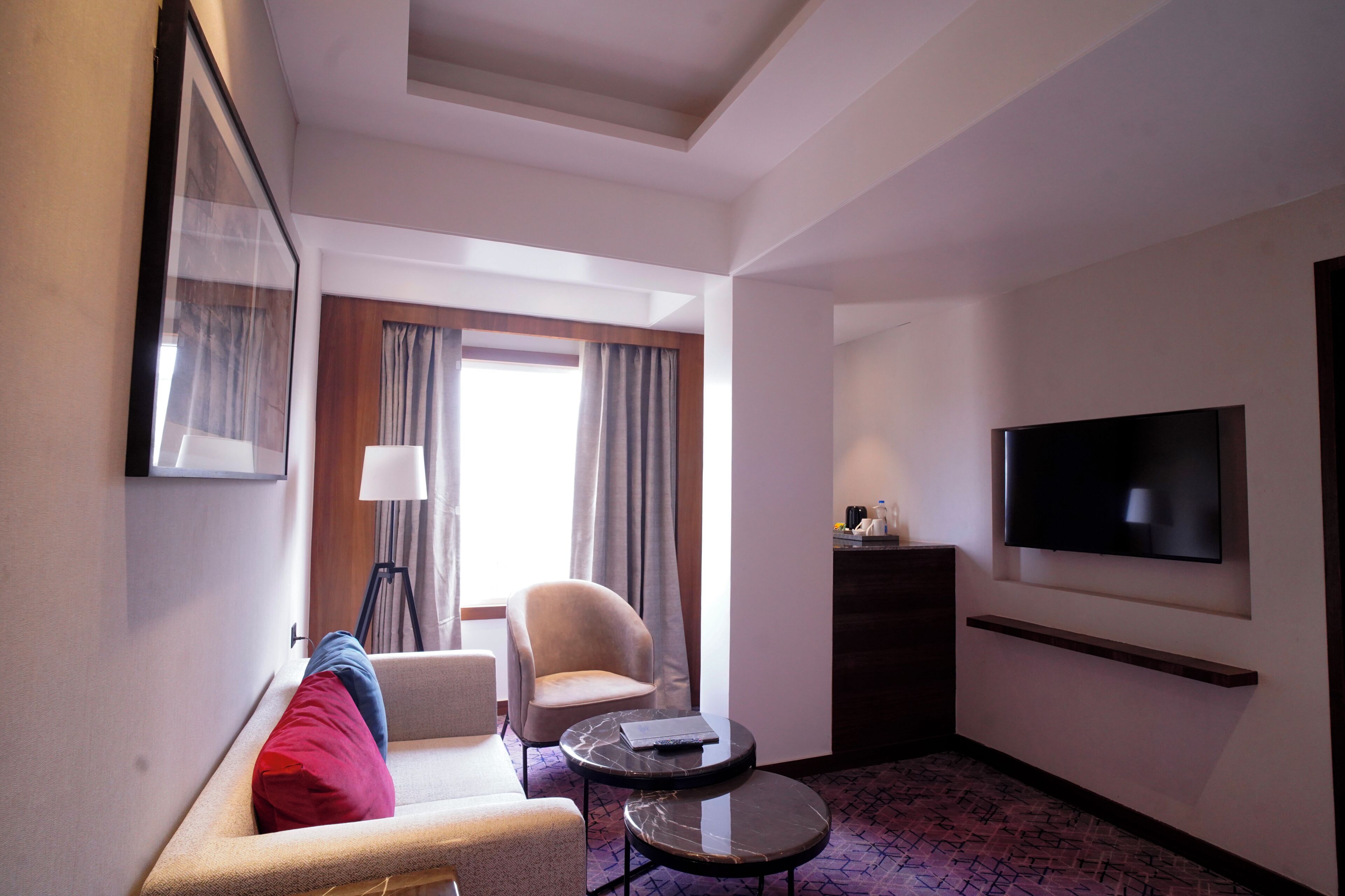 A hotel room with a sofa, armchair, small round table, TV, and a large window with curtains.