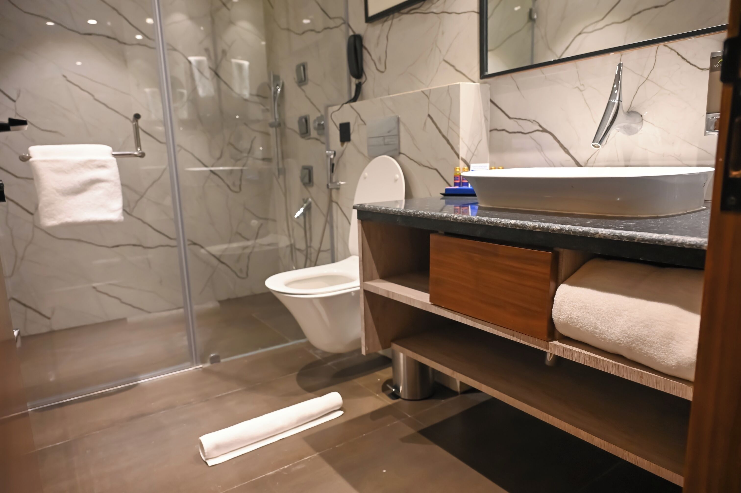 A modern bathroom with a glass shower enclosure, a white toilet, a marble-tiled wall, and a wooden vanity with a sink and towel storage.