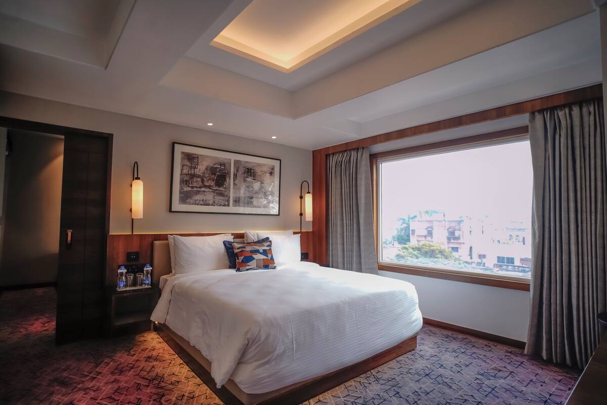 Een hotelkamer met een groot bed, een nachtkastje met een lamp, een kunstwerk in een lijst aan de muur, en een raam met gordijnen.