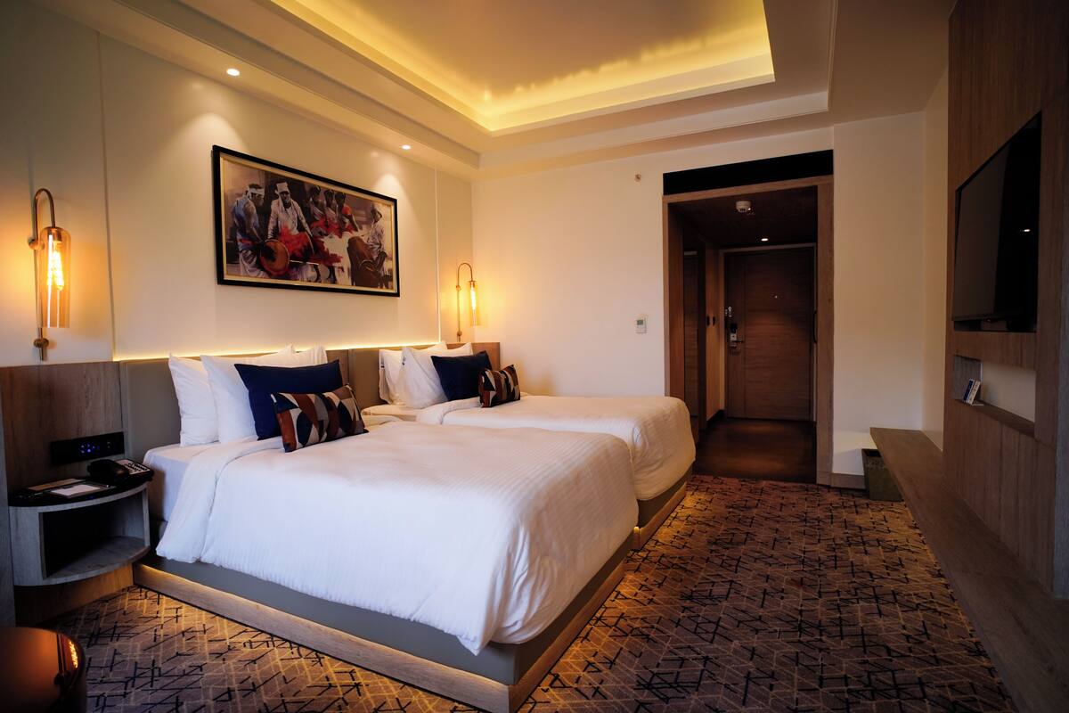 Een hotelkamer met twee bedden, een schilderij aan de muur en een aan de muur bevestigde televisie.