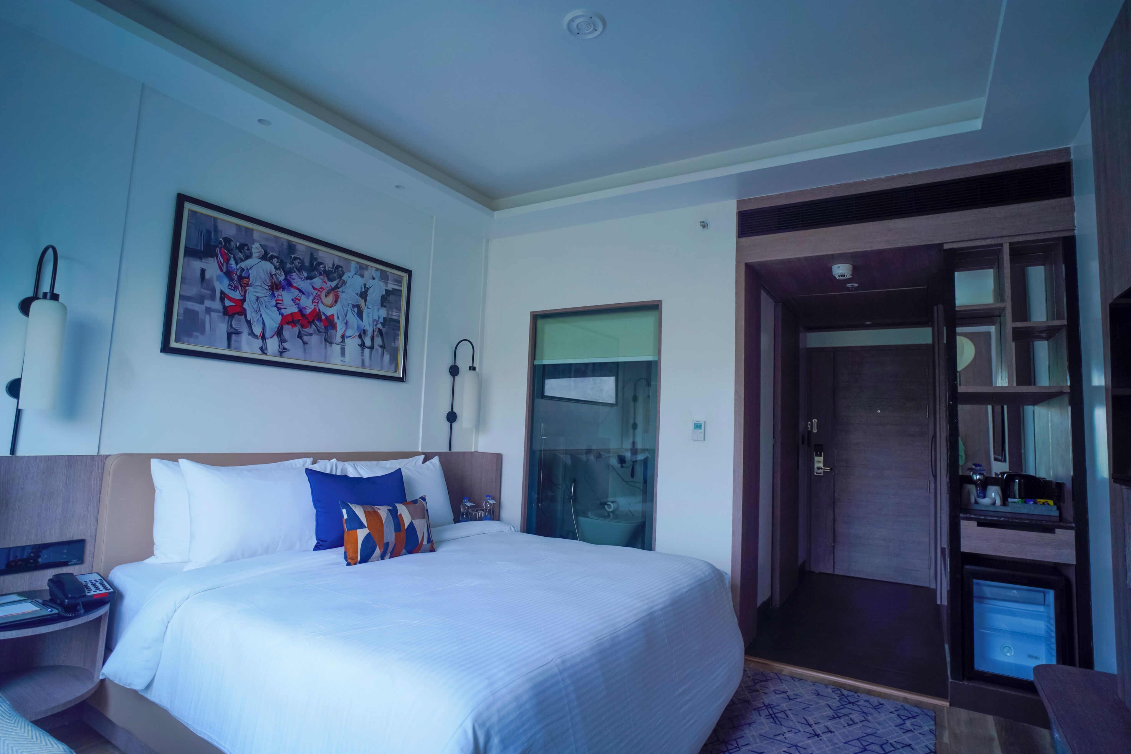 A hotel room with a large bed, a bedside table with a phone, a painting on the wall, and a door leading to a bathroom.