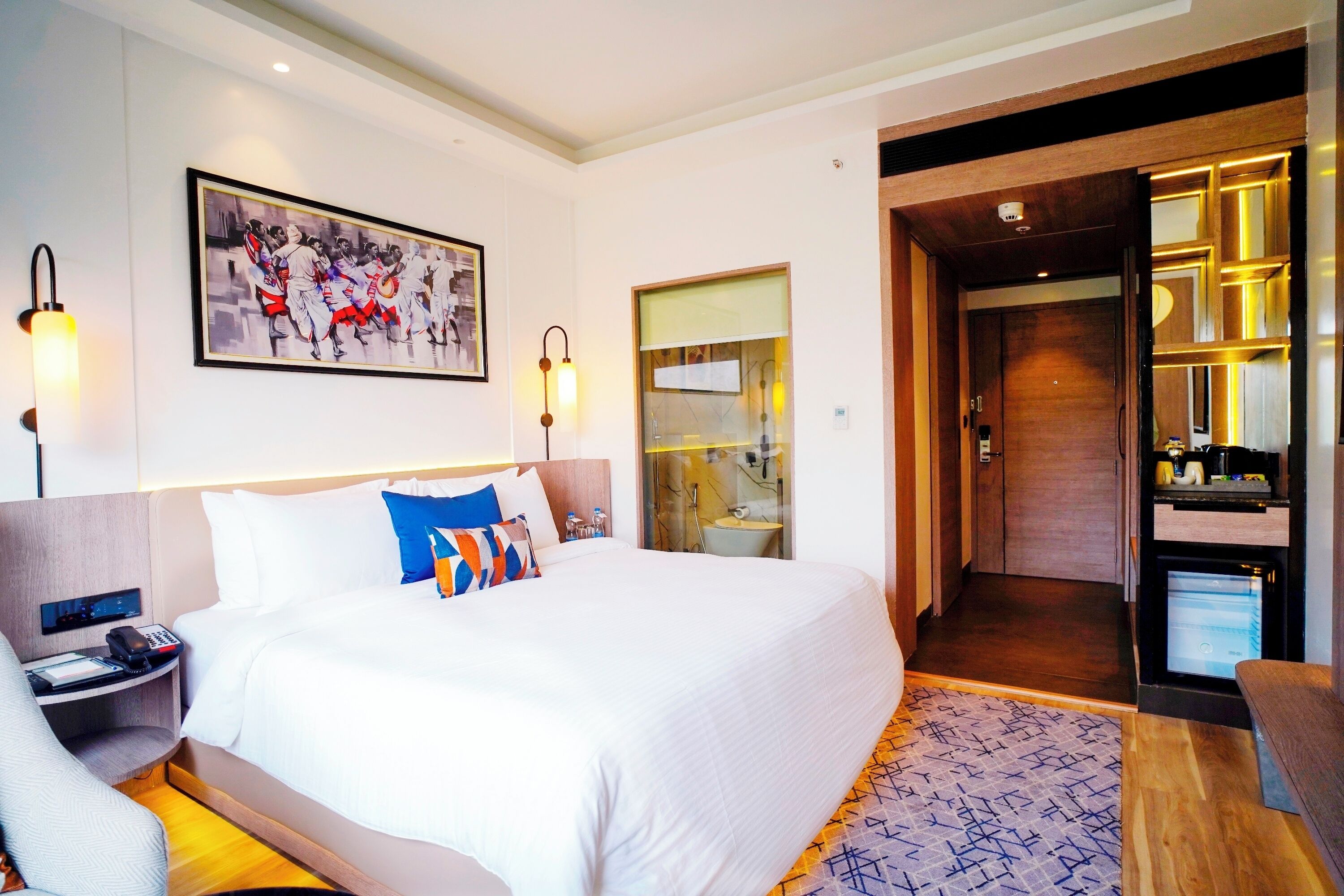 A hotel room with a large bed, a bedside table with a phone, a wall-mounted painting, and a bathroom visible through an open door.