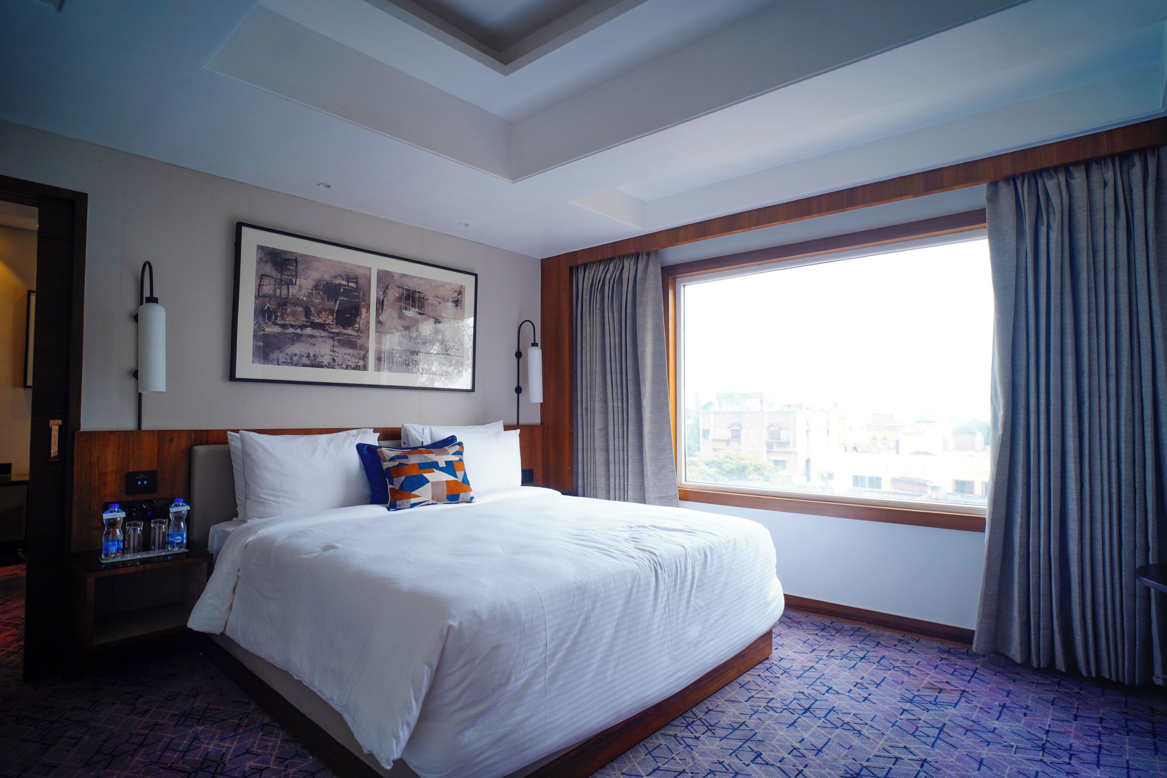 A hotel room with a large bed, a nightstand with a lamp, a framed picture on the wall, and a window with curtains.