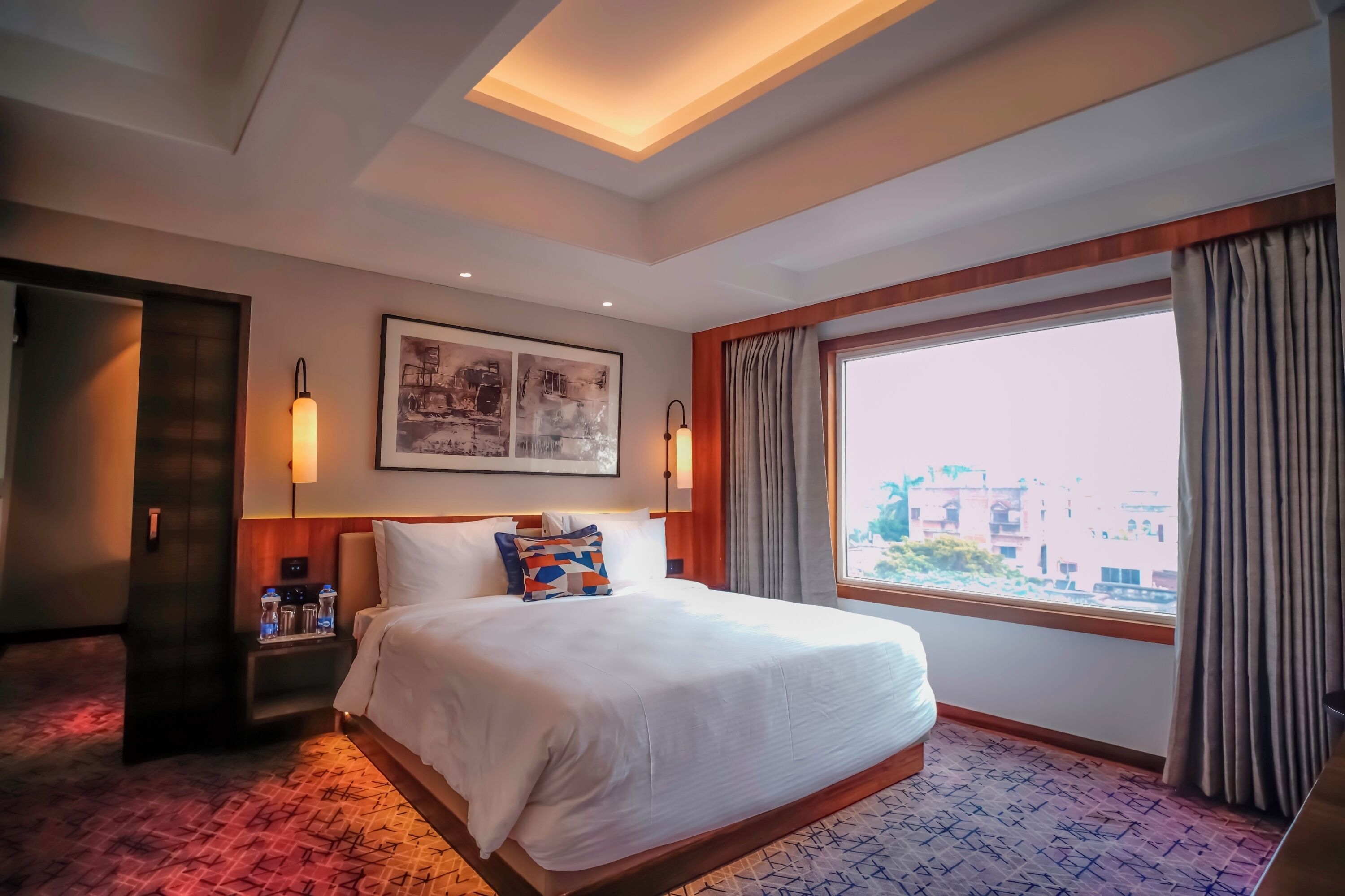 Een hotelkamer met een groot bed, een nachtkastje met lamp, een kunstwerk in een lijst aan de muur en een raam met gordijnen.