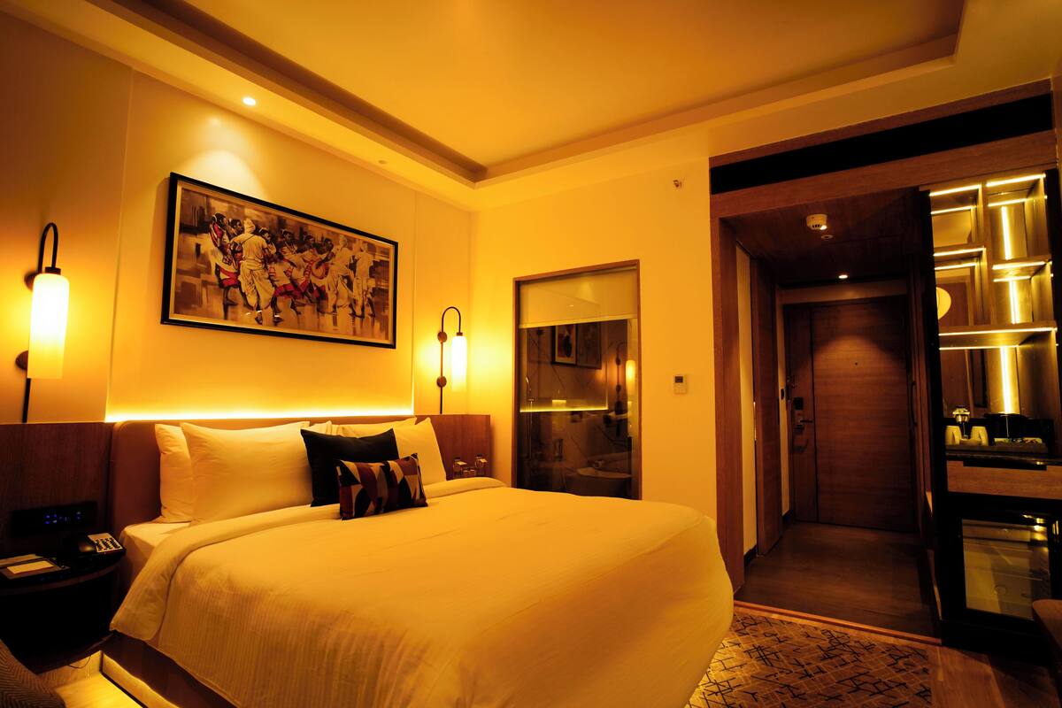 Een hotelkamer met een groot bed, bedlampjes, een schilderij aan de muur, een spiegel en een deur naar een badkamer.