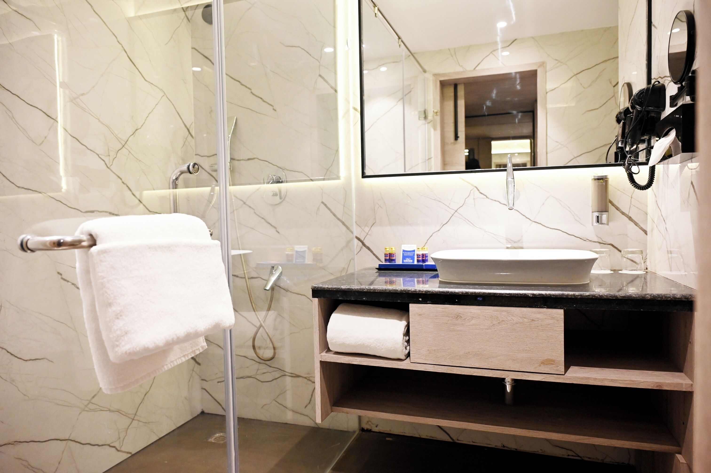 Un baño moderno con cabina de ducha de vidrio, pared revestida de mármol, lavamanos blanco y tocador de madera.