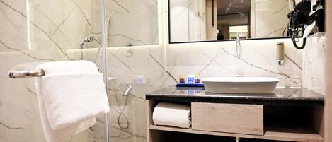 Un baño moderno con cabina de ducha de vidrio, pared revestida de mármol, lavamanos blanco y tocador de madera.