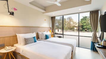 Habitación de hotel con dos camas, ventilador de techo, aire acondicionado, escritorio con lámpara y ventana grande con vistas a árboles y edificios.