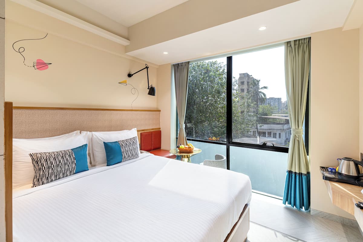 Ein Hotelzimmer mit einem großen Bett, einem kleinen Tisch mit Obstschale, einem Schreibtisch mit Wasserkocher und Blick auf die Stadt durch ein großes Fenster.