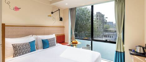 A hotel room with a large bed, a small table with a fruit bowl, a desk with a kettle, and a view of the city through a large window.