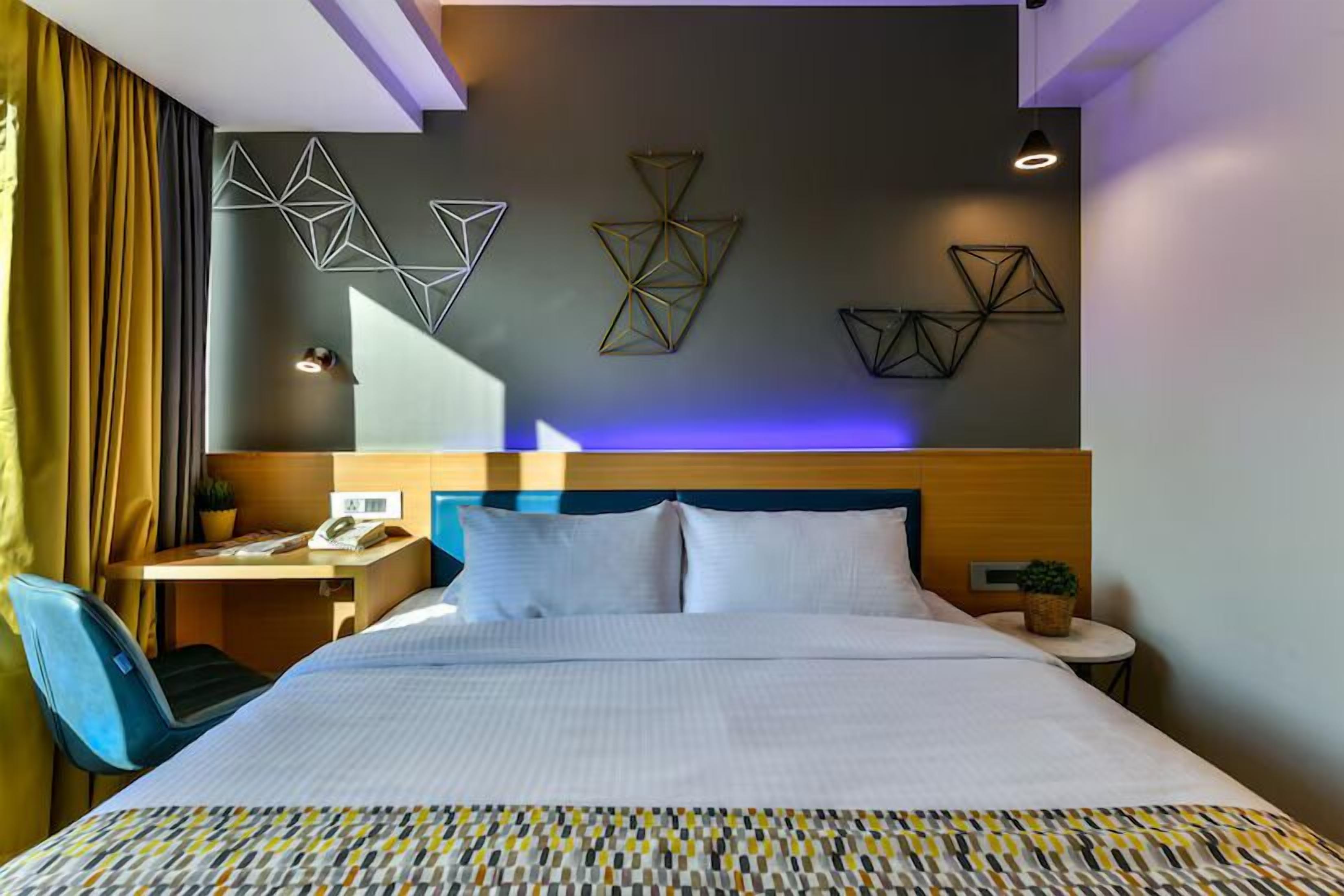 Una habitación de hotel con una cama grande, un escritorio con una silla, una mesita con una planta y arte geométrico en la pared.