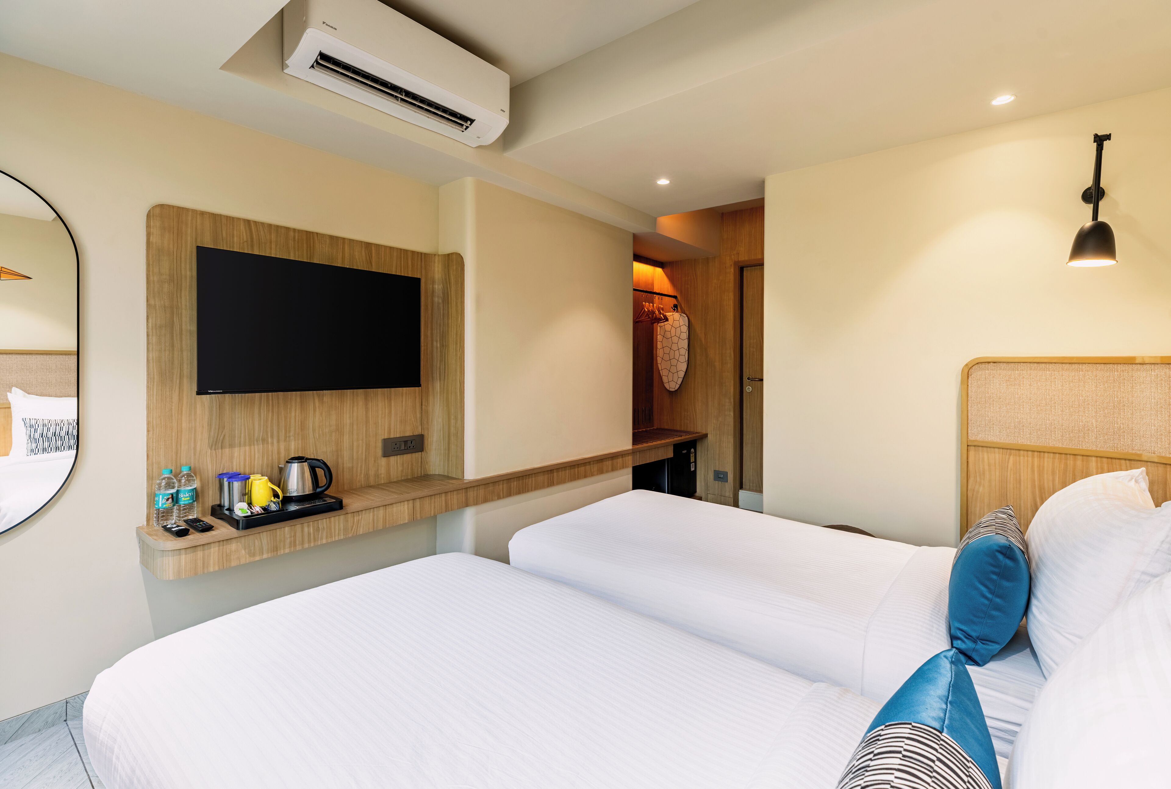 Habitación de hotel con dos camas, un televisor de pantalla plana montado en la pared, un pequeño estante con artículos de tocador y un armario con un bolso colgado.
