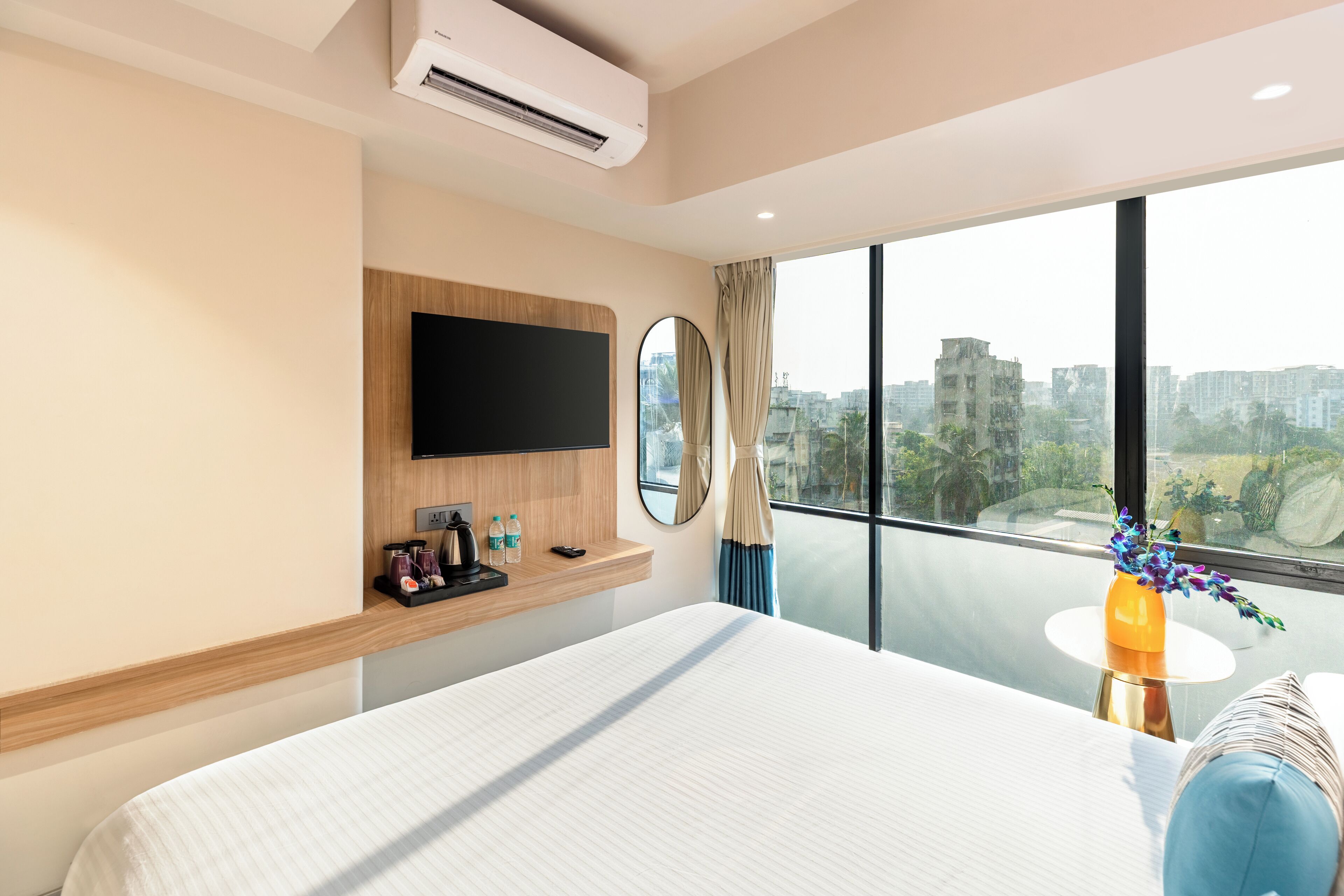 Habitación de hotel con una cama grande, un televisor montado, una mesita de noche con un florero y vistas a la ciudad a través de amplios ventanales.